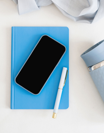 a flat lay image of a deep sky blue notebook with an mobile phone on top and a white pen plus a light blue tumbler