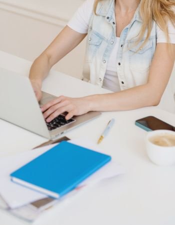 a woman typing on a laptop with a sky blue notebook, a cup of coffee and a mobile phone atop her desk