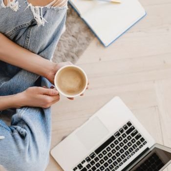 an image of a woman in jeans sitting on the floor holding a cup of coffee while looking down on an open laptop with an open notebook on her left